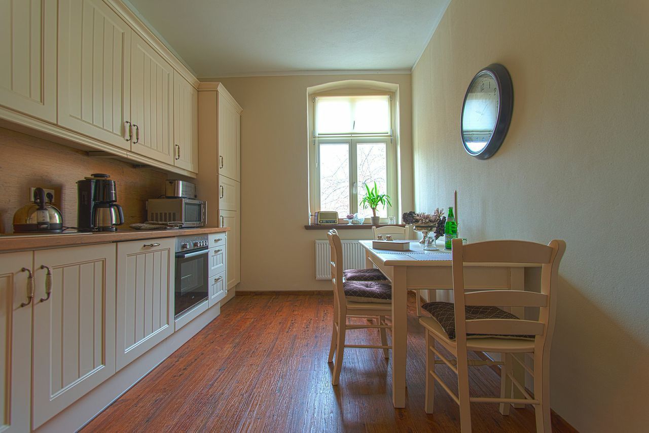 A kitchen with wooden cabinets, a dining table with chairs, a window with a view, and a wall clock.