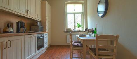 A kitchen with wooden cabinets, a dining table with chairs, a window with a view, and a wall clock.