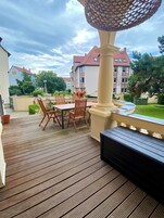 Una terraza de madera con una mesa y sillas, una lámpara decorativa y vistas a edificios al fondo.