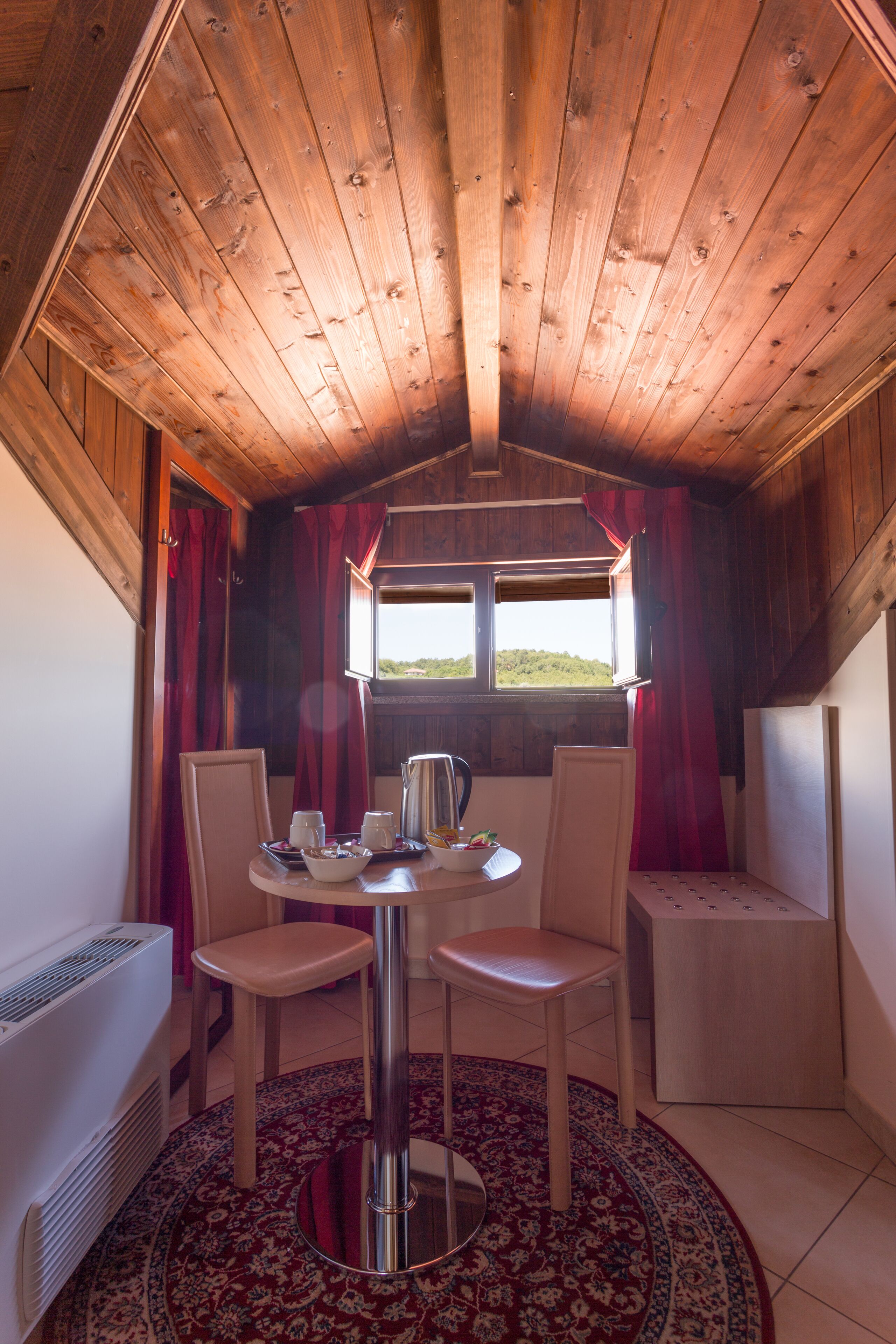 Une pièce chaleureuse avec un plafond en bois, une table ronde entourée de chaises, une fenêtre avec des rideaux et un radiateur.