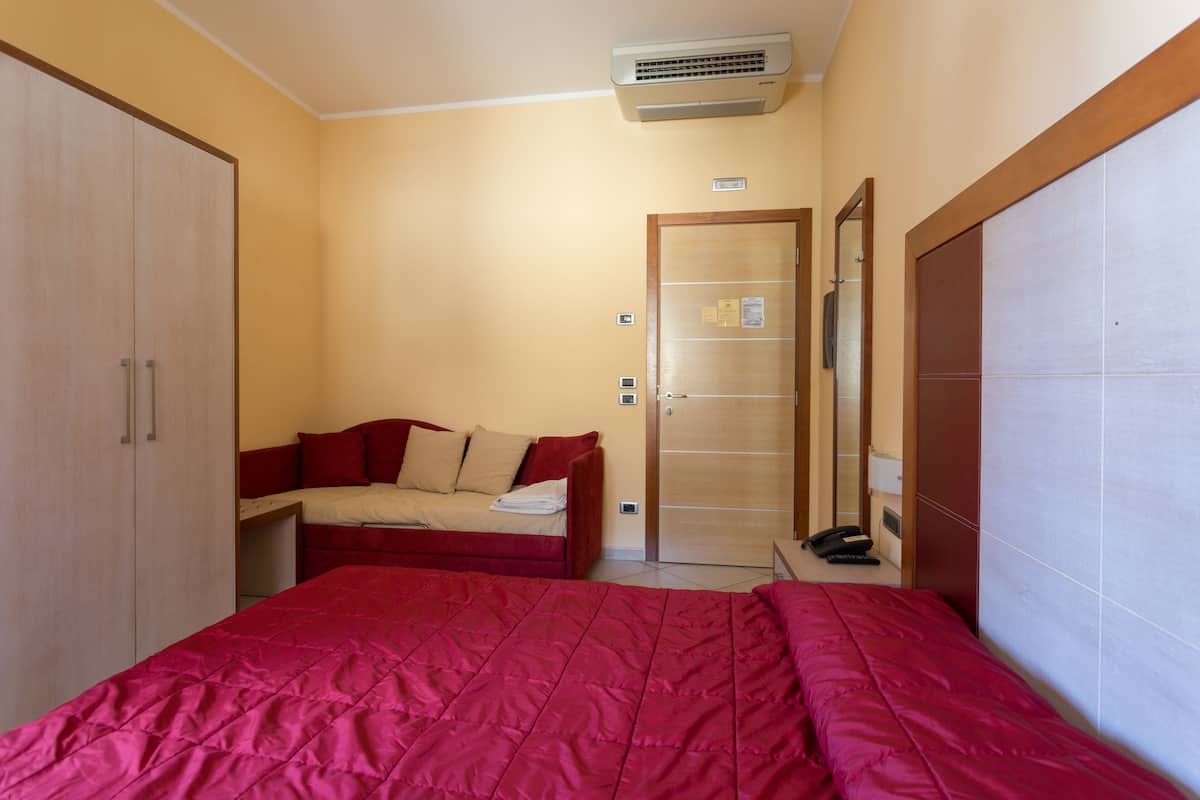 A bedroom with a large bed, a small sofa, a wardrobe, a mirror, and an air conditioning unit.