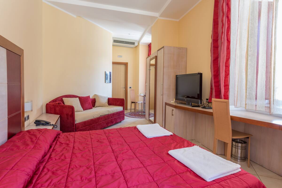 A hotel room with a large bed, a sofa, a desk with a TV, and a window with curtains.