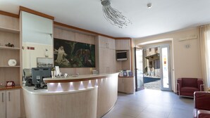 A reception area with a curved counter, a large painting, a television, and a seating area with red chairs. - Hotel Conte Ruggero (Serra San Bruno)