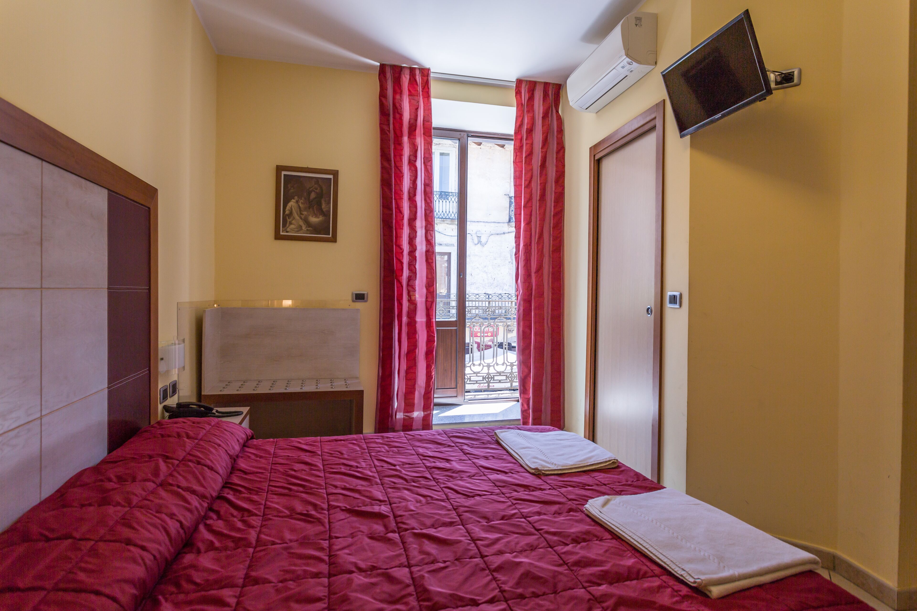 Une chambre d’hôtel avec un lit, un téléviseur à écran plat fixé au mur, une fenêtre avec des rideaux rouges et un balcon visible à travers la fenêtre.