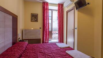 Une chambre d’hôtel avec un lit, un téléviseur à écran plat fixé au mur, une fenêtre avec des rideaux rouges et un balcon visible à travers la fenêtre.