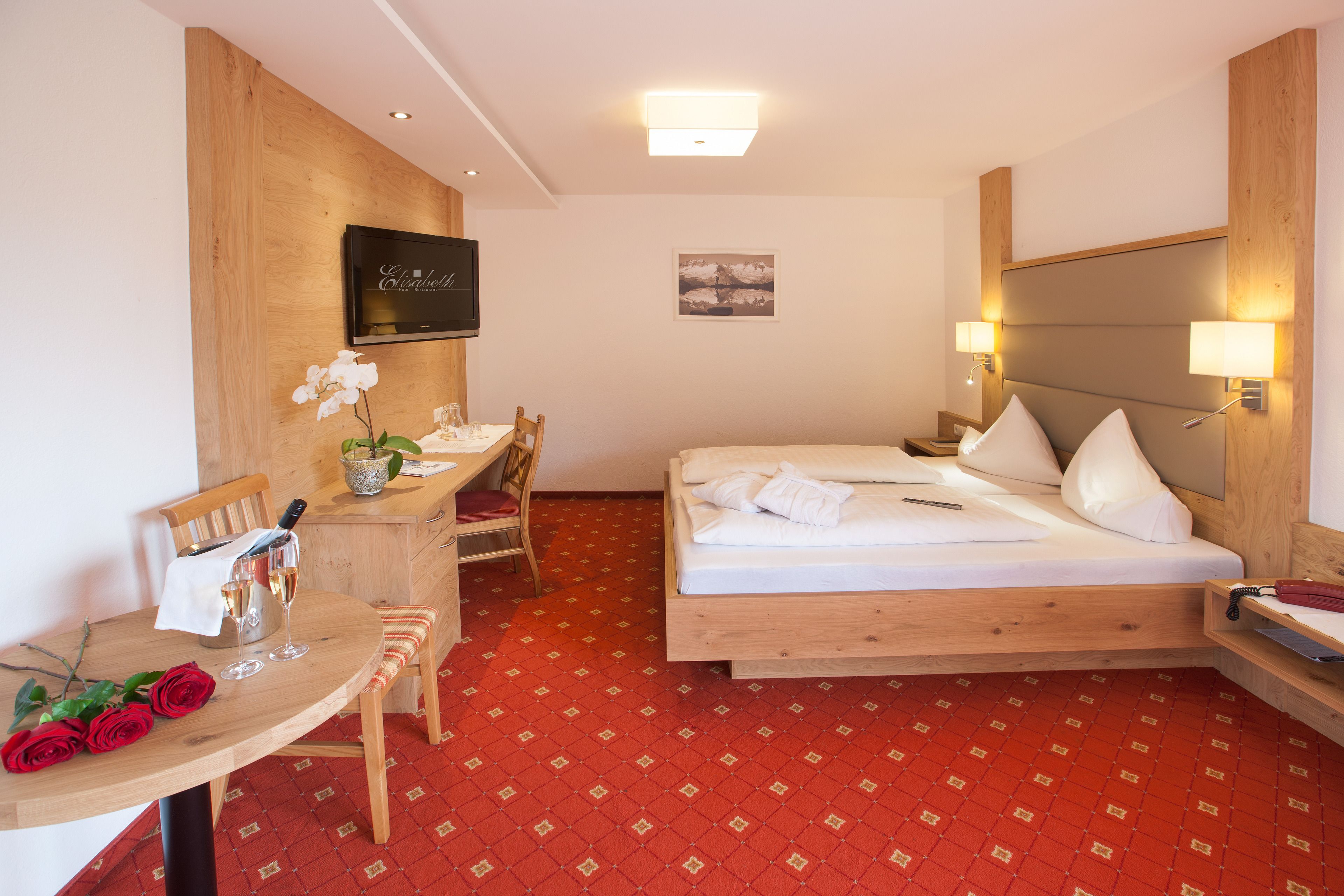 Habitación de hotel con cabecera de madera, cama con ropa blanca, escritorio con silla, una mesita con un florero y una alfombra roja estampada.
