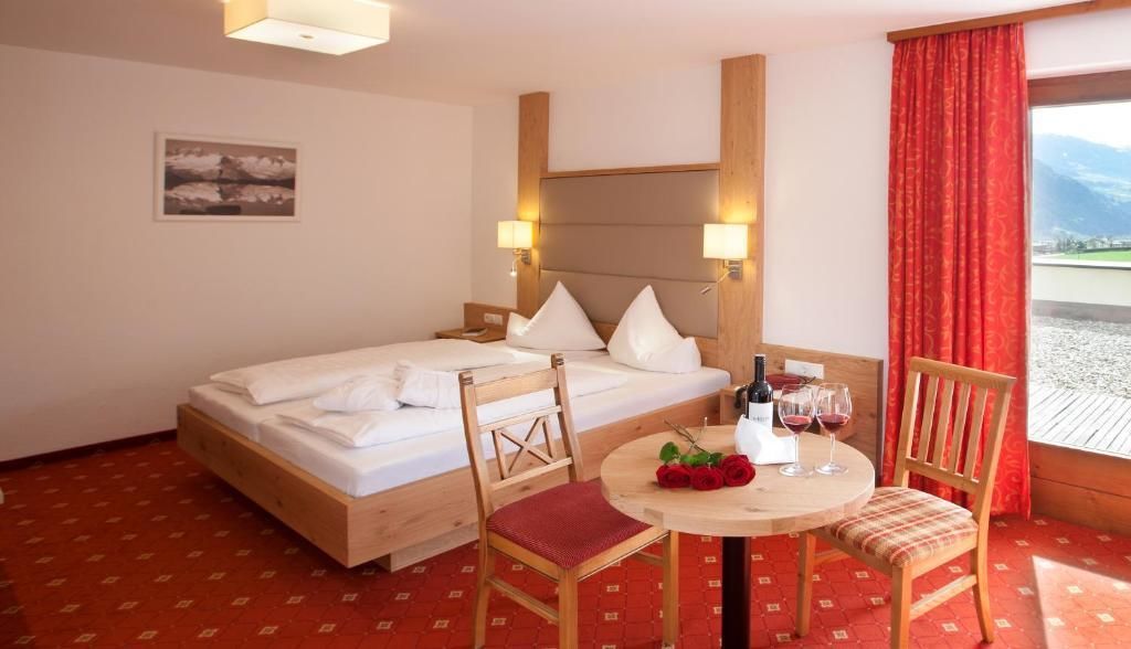 A hotel room with a bed, two chairs, a small table with wine glasses and a bouquet of roses, and a window with a red curtain.