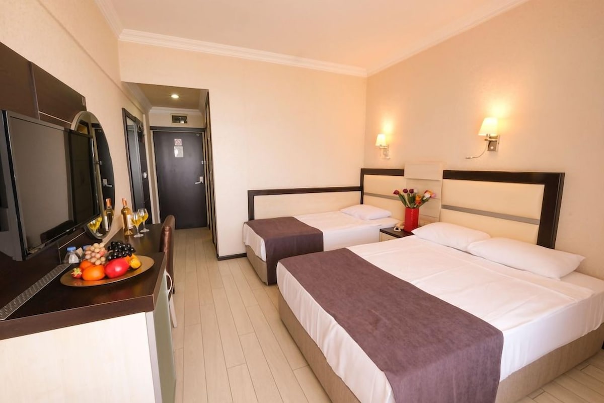 Habitación de hotel con dos camas, un escritorio con una bandeja de frutas, un televisor y un espejo.