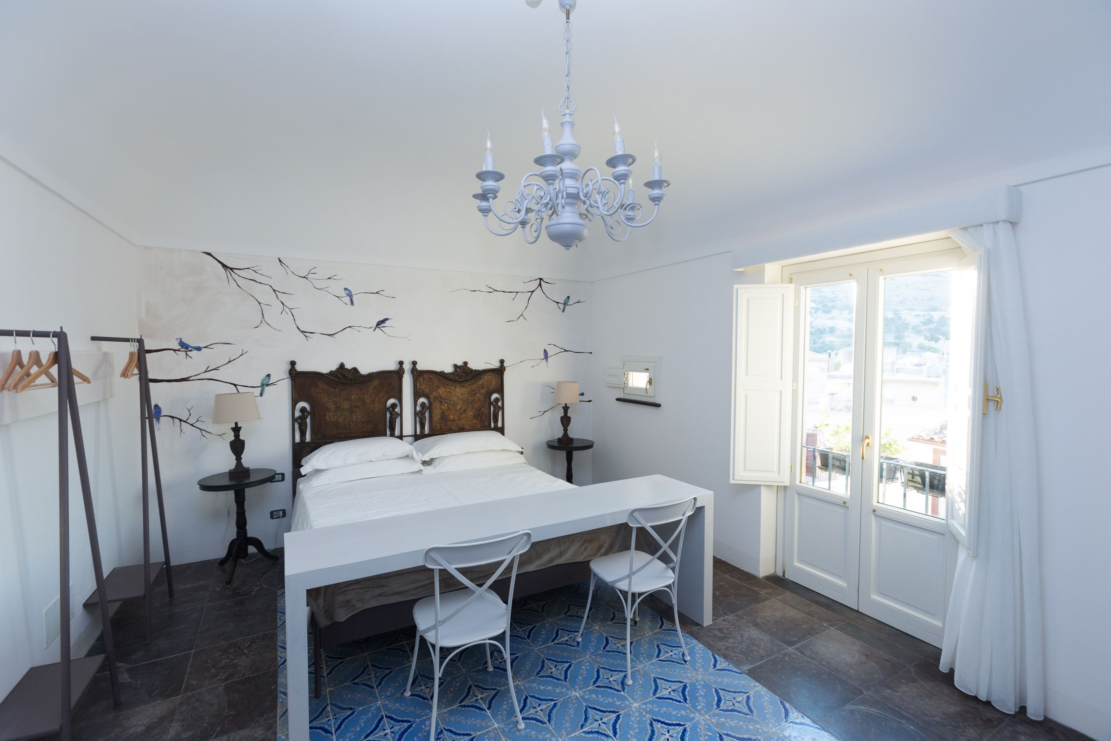 A bedroom with a bed, two chairs, a chandelier, a window, and wall art.