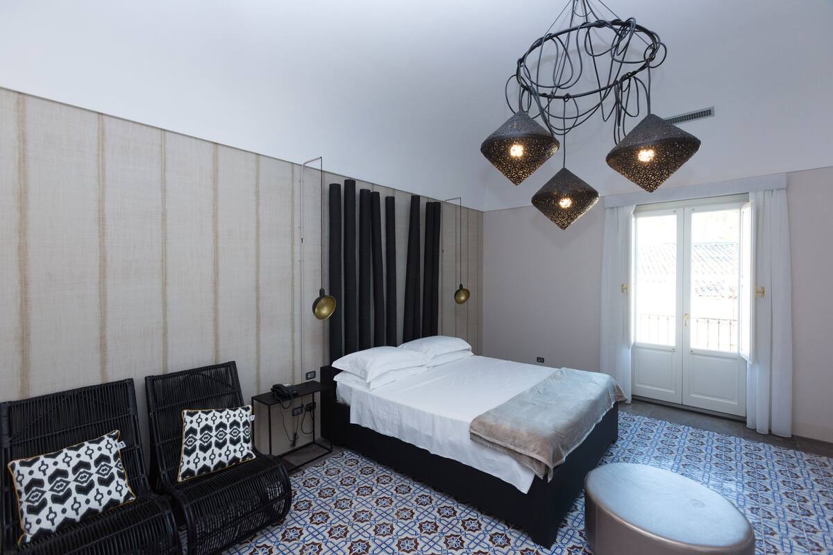 A bedroom with a bed, two bedside tables, a chair, a patterned rug, and a unique chandelier.