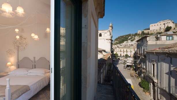 A bedroom with a bed, bedside tables, and hanging lights. The room has a large window with a view of a street and buildings. - Palazzo Conti Camere & Suite (Scicli)