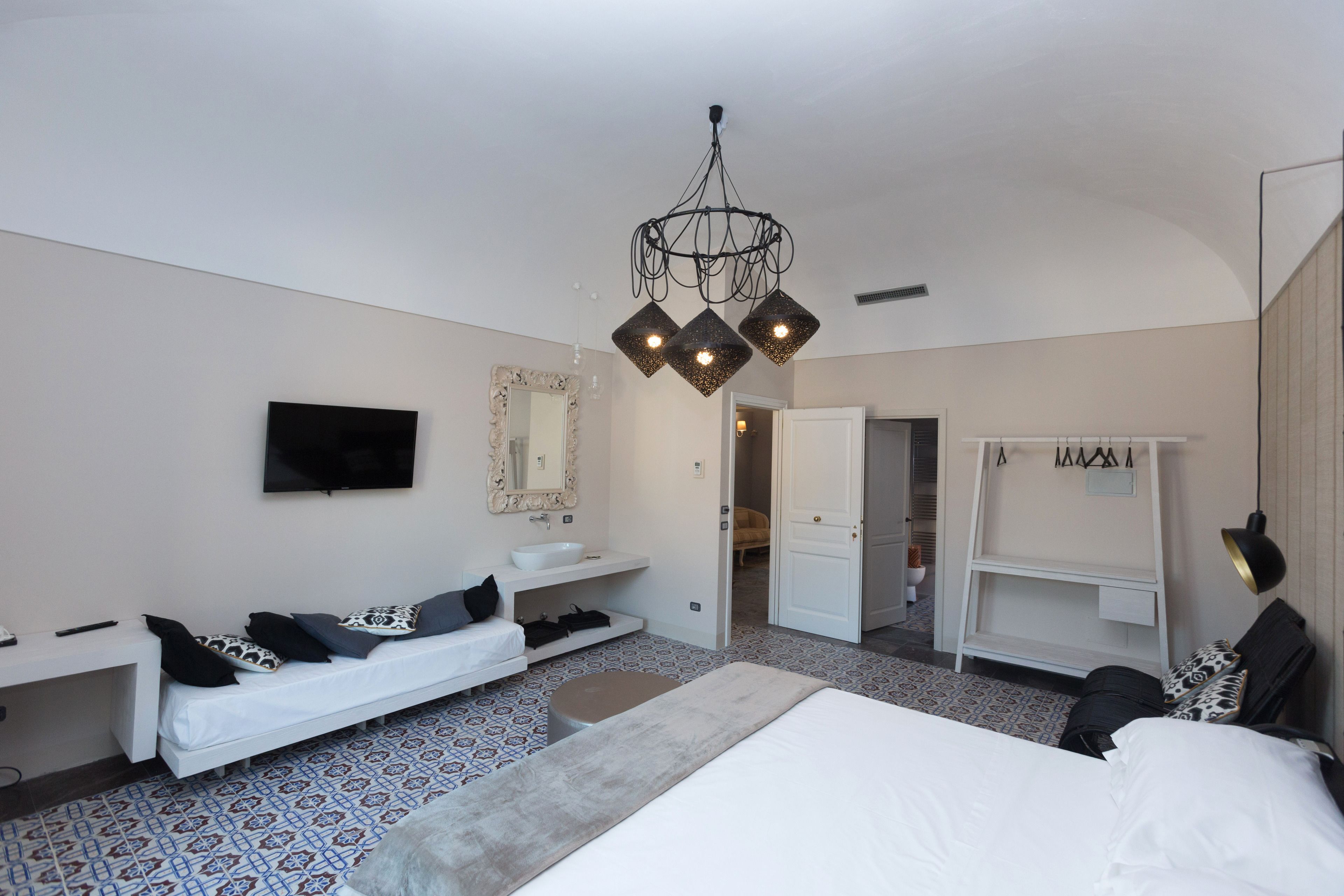 A modern hotel room with a large bed, a sofa, a vanity with a mirror, a television, and a chandelier.