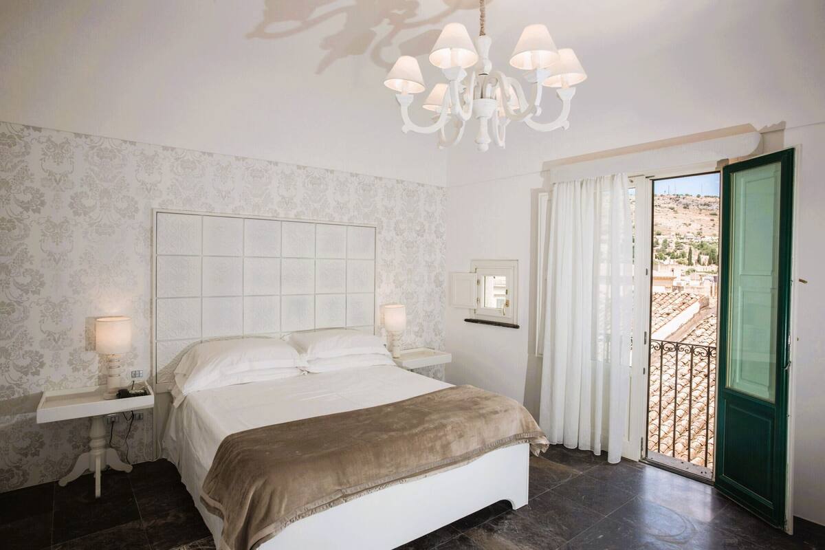 A bedroom with a large bed, a chandelier, a nightstand, a window with curtains, and a view of a landscape.
