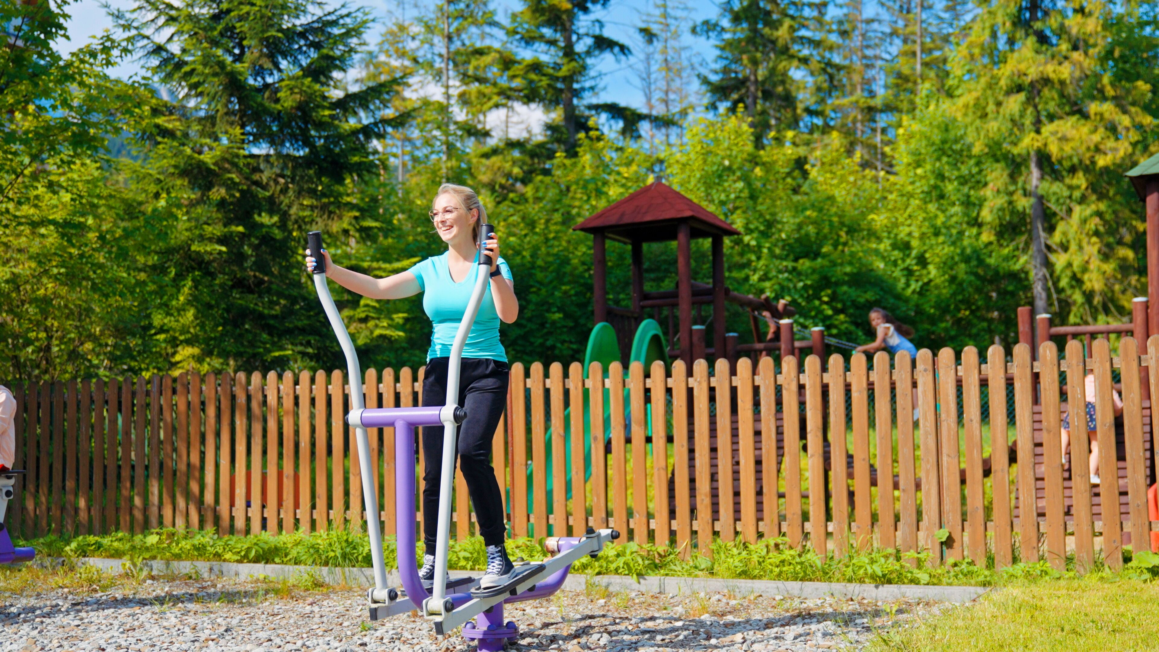 Una persona usando un equipo de ejercicio al aire libre en un parque con juegos infantiles y una valla de madera al fondo.