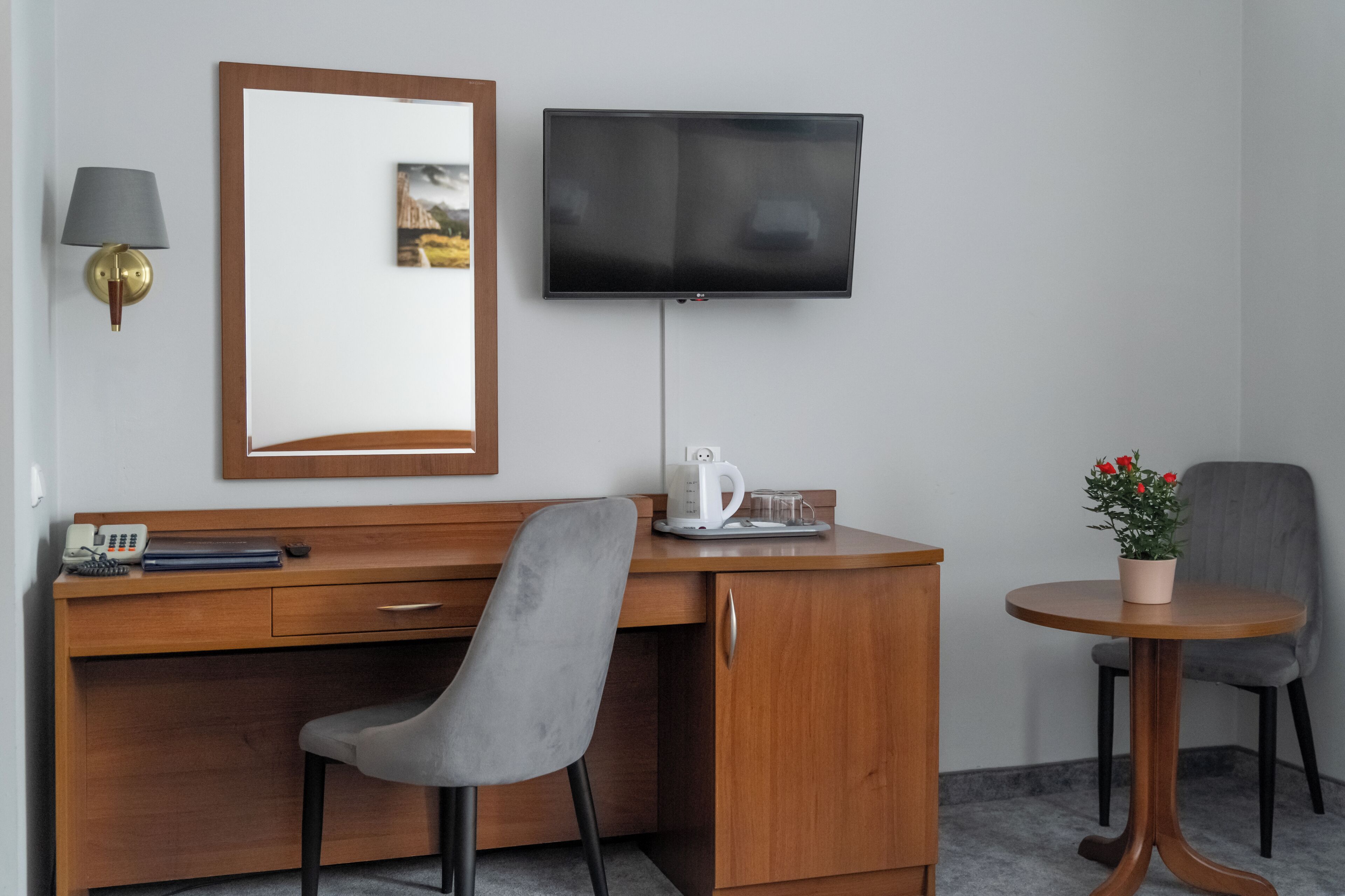 Ein Hotelzimmer mit einem Holztisch, einem grauen Sessel, einem an der Wand befestigten Fernseher, einem kleinen Tisch mit Kaffeemaschine und einer Topfpflanze.