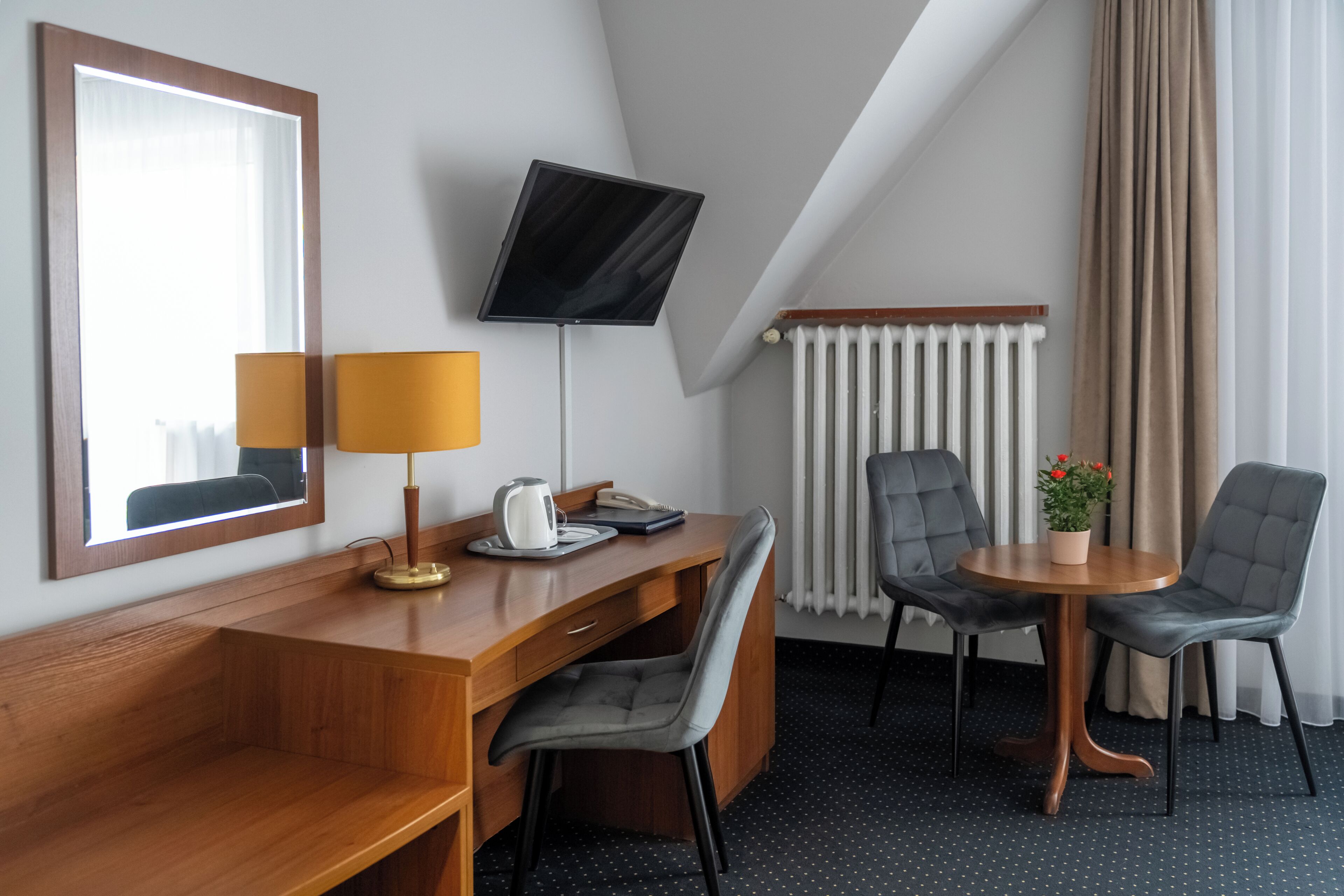 A hotel room with a wooden desk, two chairs, a television, a lamp, a radiator, and a potted plant.