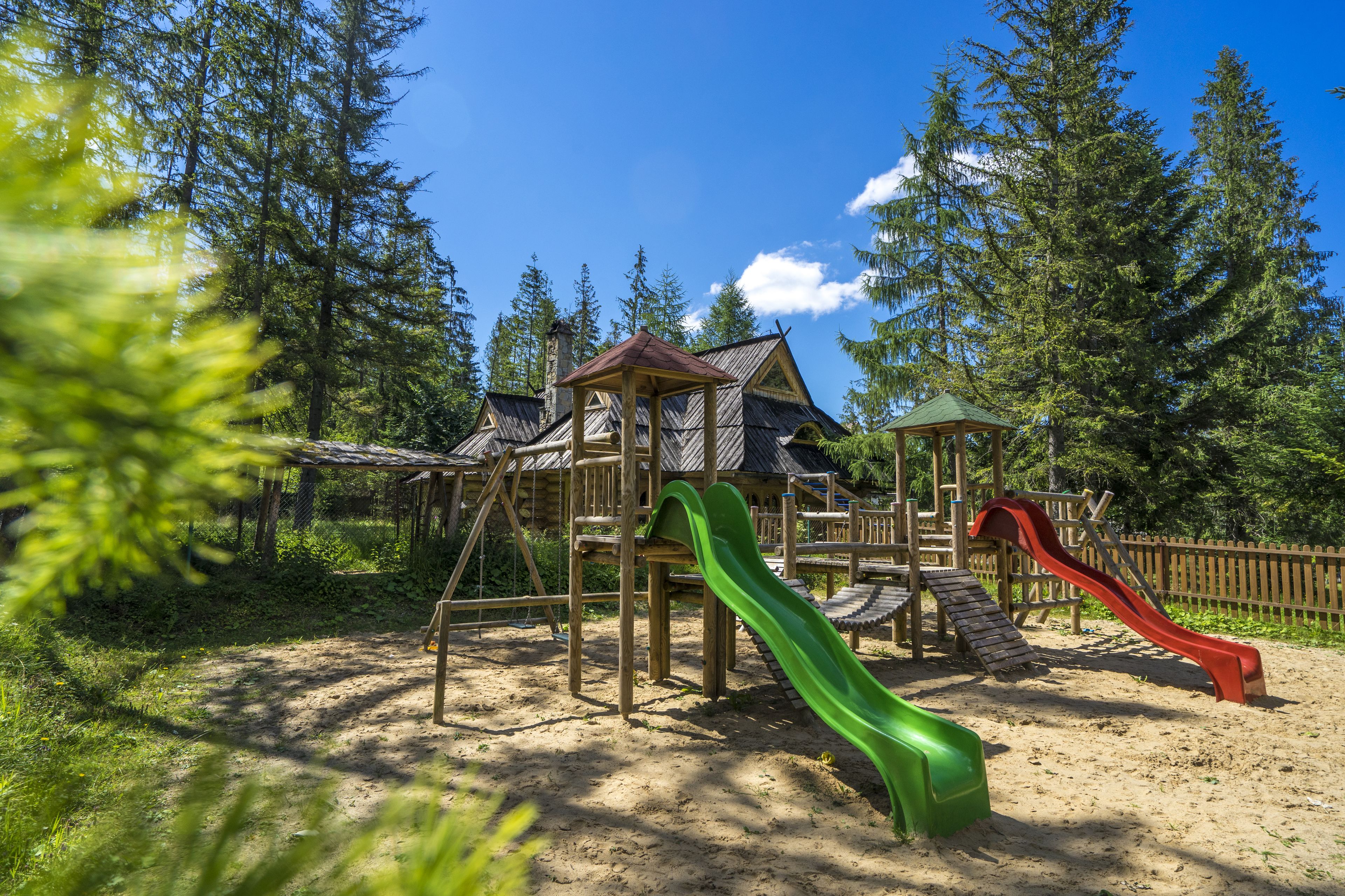 A wooden play structure with two slides and a small building in a forested area.