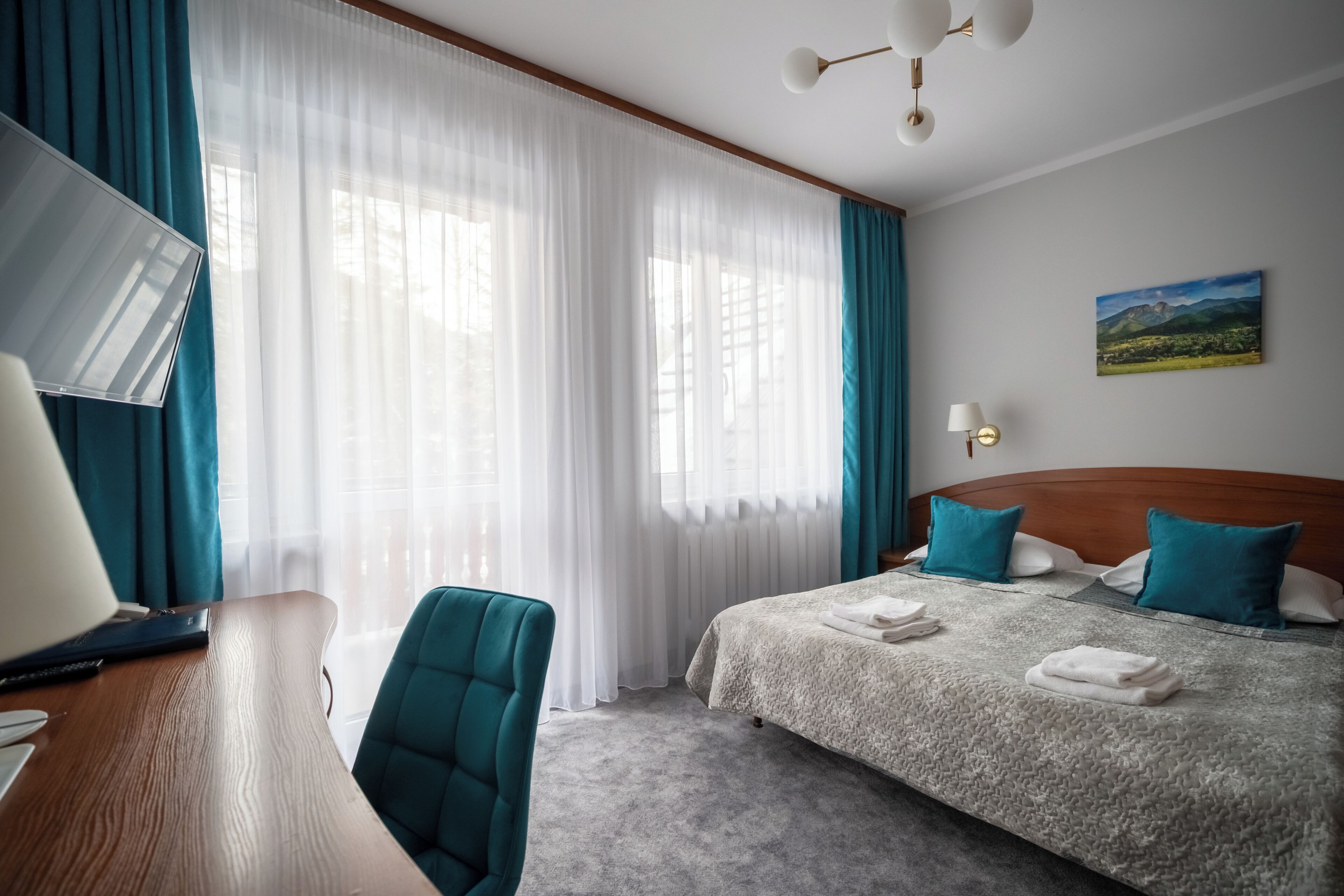 A hotel room with a bed, a desk, a chair, a TV, and a painting on the wall.