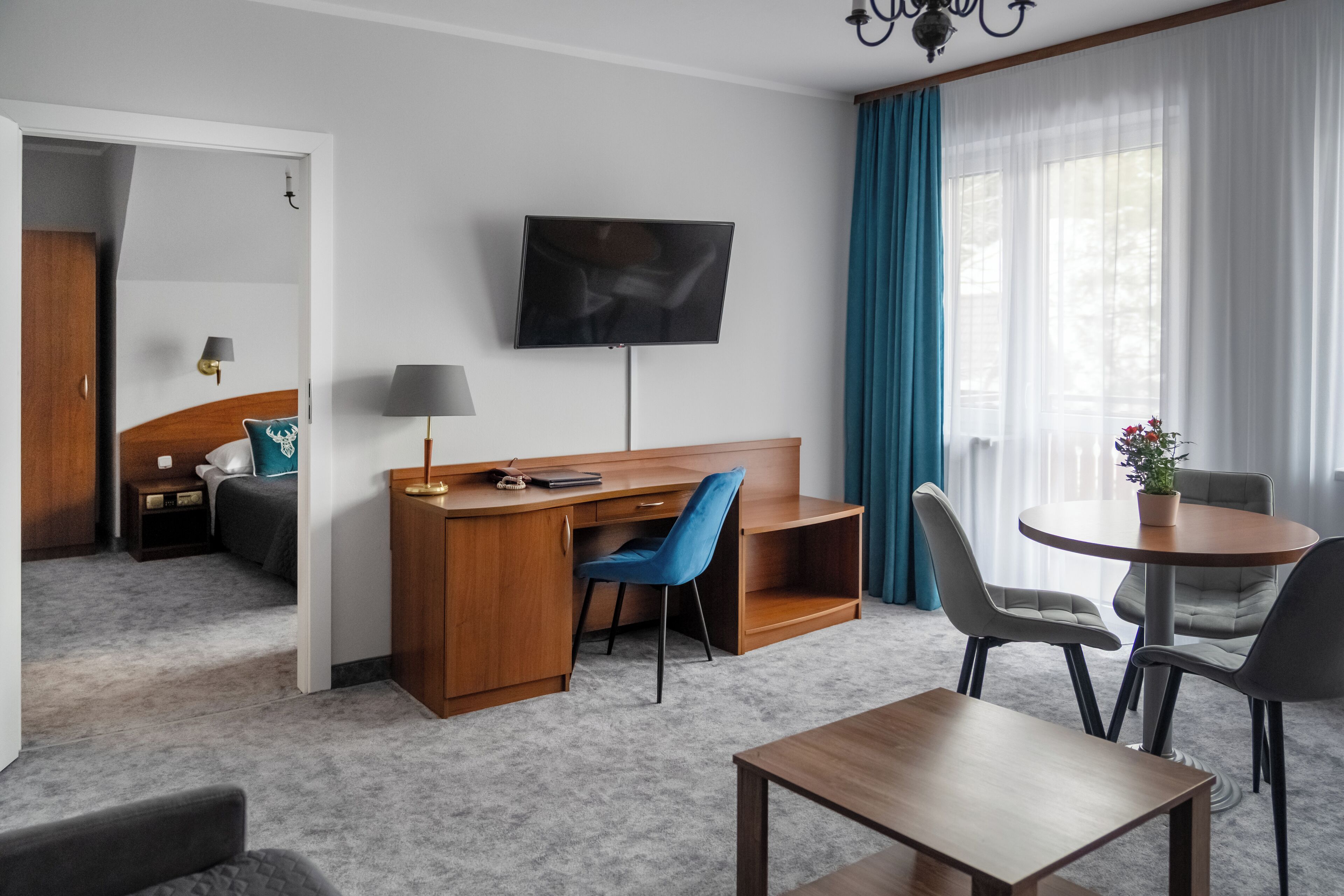 A hotel room with a wooden desk, a blue chair, a television, a small table, and a dining area with chairs.