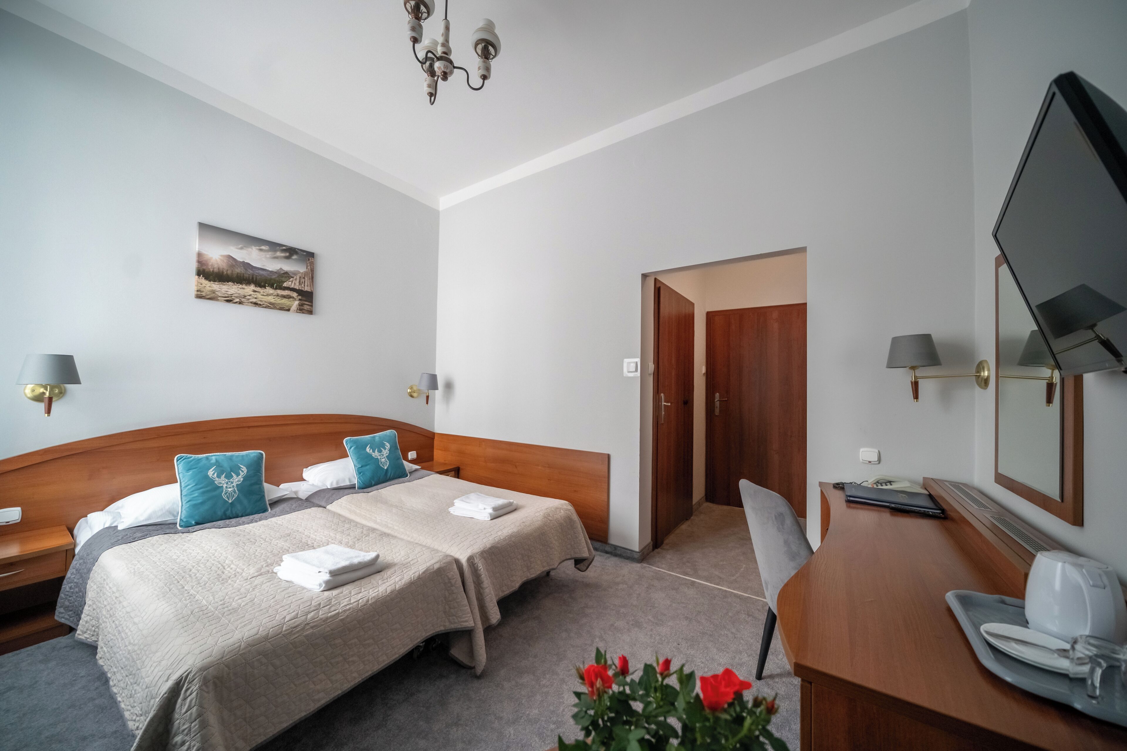 A hotel room with two beds, a desk, a chair, a desk lamp, a telephone, a mirror, and a painting on the wall.