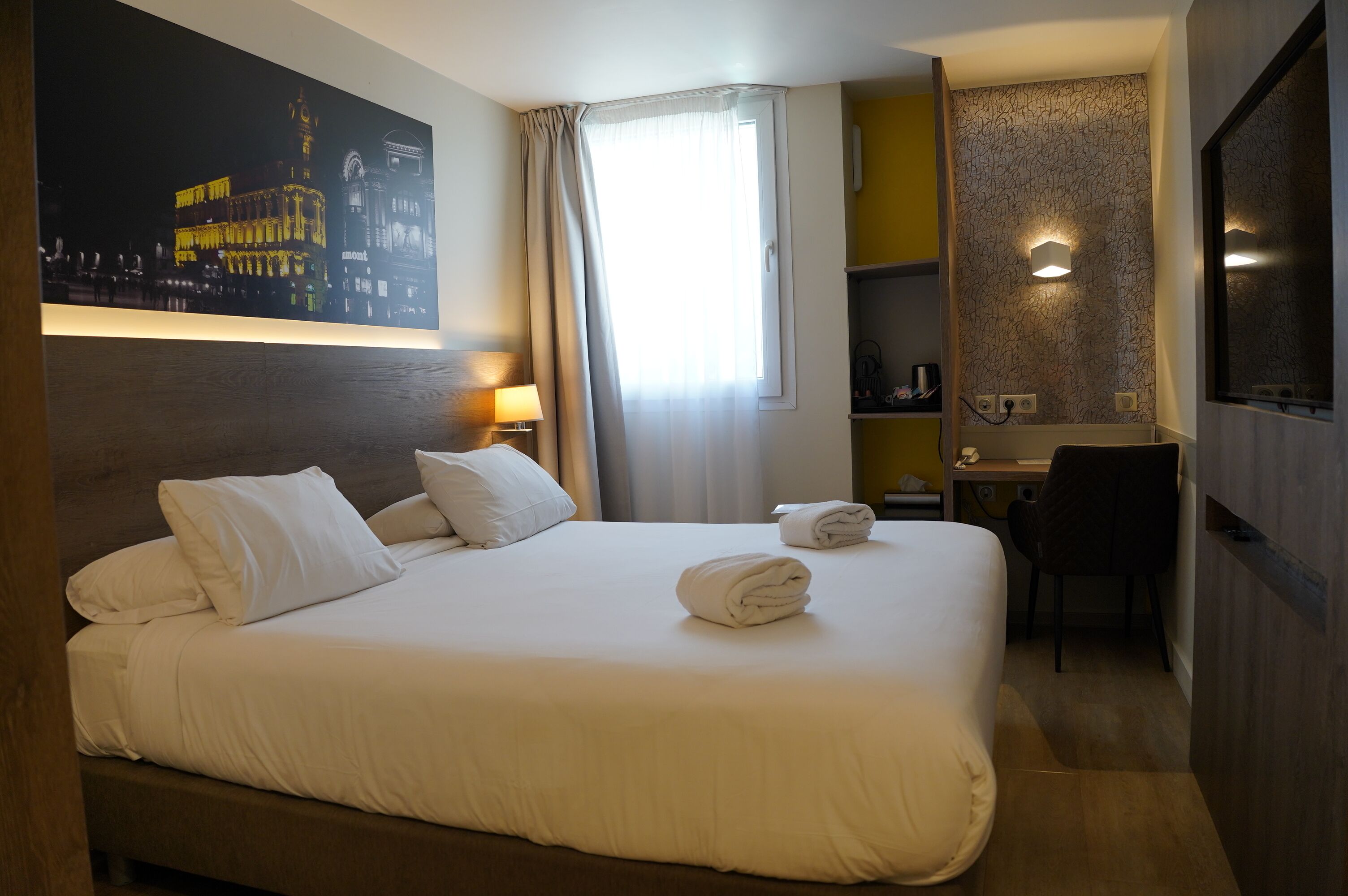 Una habitación de hotel con cama, escritorio, silla, un cuadro y ventana con cortinas.