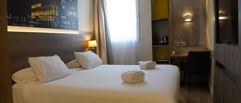 A hotel room with a bed, a desk, a chair, a painting, and a window with curtains.