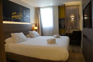 Una habitación de hotel con cama, escritorio, silla, un cuadro y ventana con cortinas.
