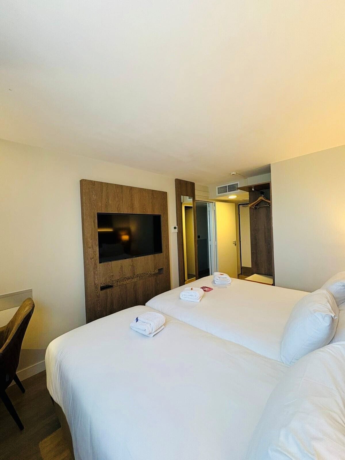 Une chambre d’hôtel avec deux lits, un téléviseur fixé sur un panneau en bois, une armoire et un miroir.
