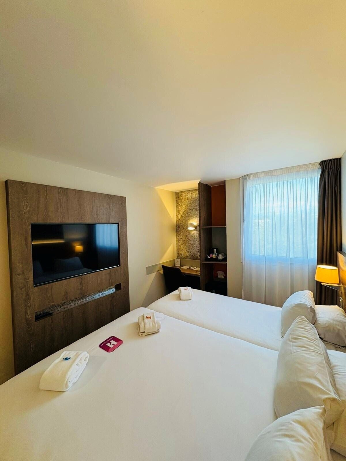 A hotel room with two beds, a flat-screen TV, a desk, a lamp, and a window with curtains.