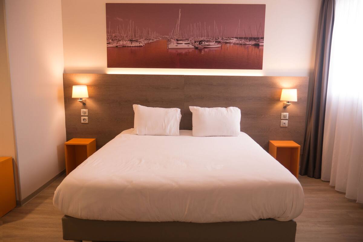 Habitación de hotel con una cama grande, dos lámparas de noche, una obra de arte en la pared que representa veleros y suelo de madera.