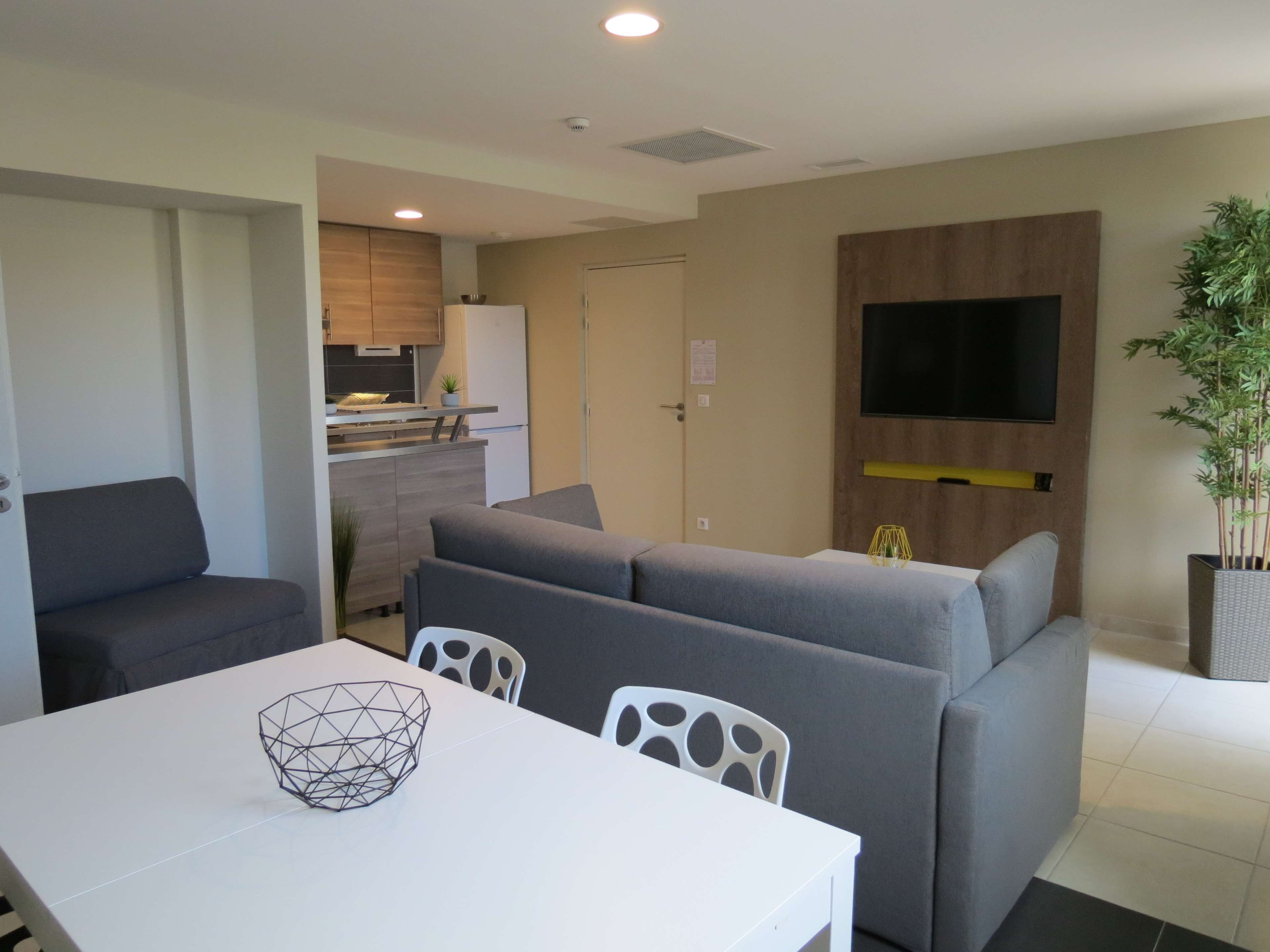 A modern living room with a grey sofa, a white dining table, and a TV mounted on the wall.