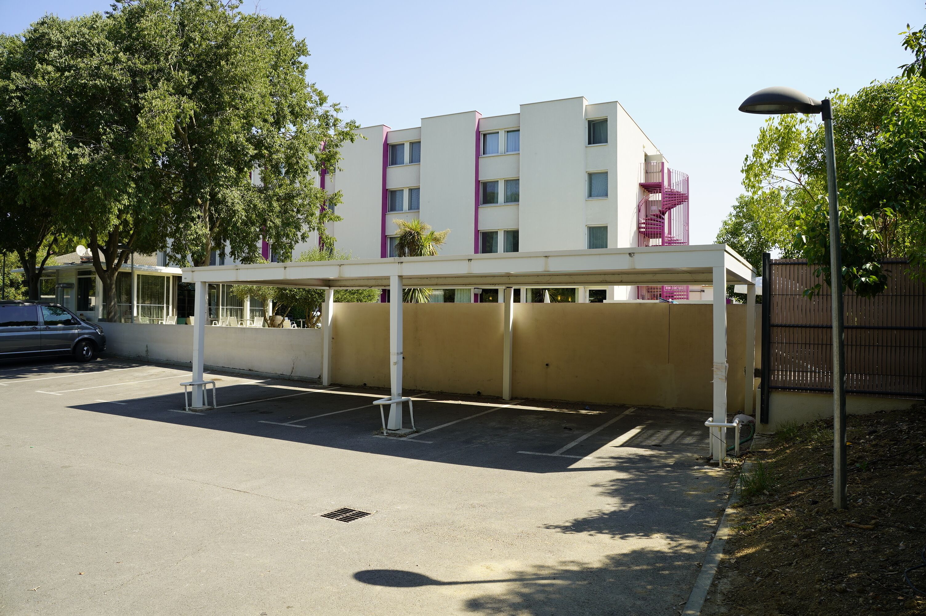 Un espace de stationnement doté d’une zone abritée, un bâtiment avec un escalier violet et un arbre.
