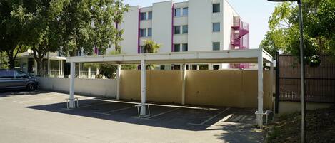 Un espace de stationnement doté d’une zone abritée, un bâtiment avec un escalier violet et un arbre.