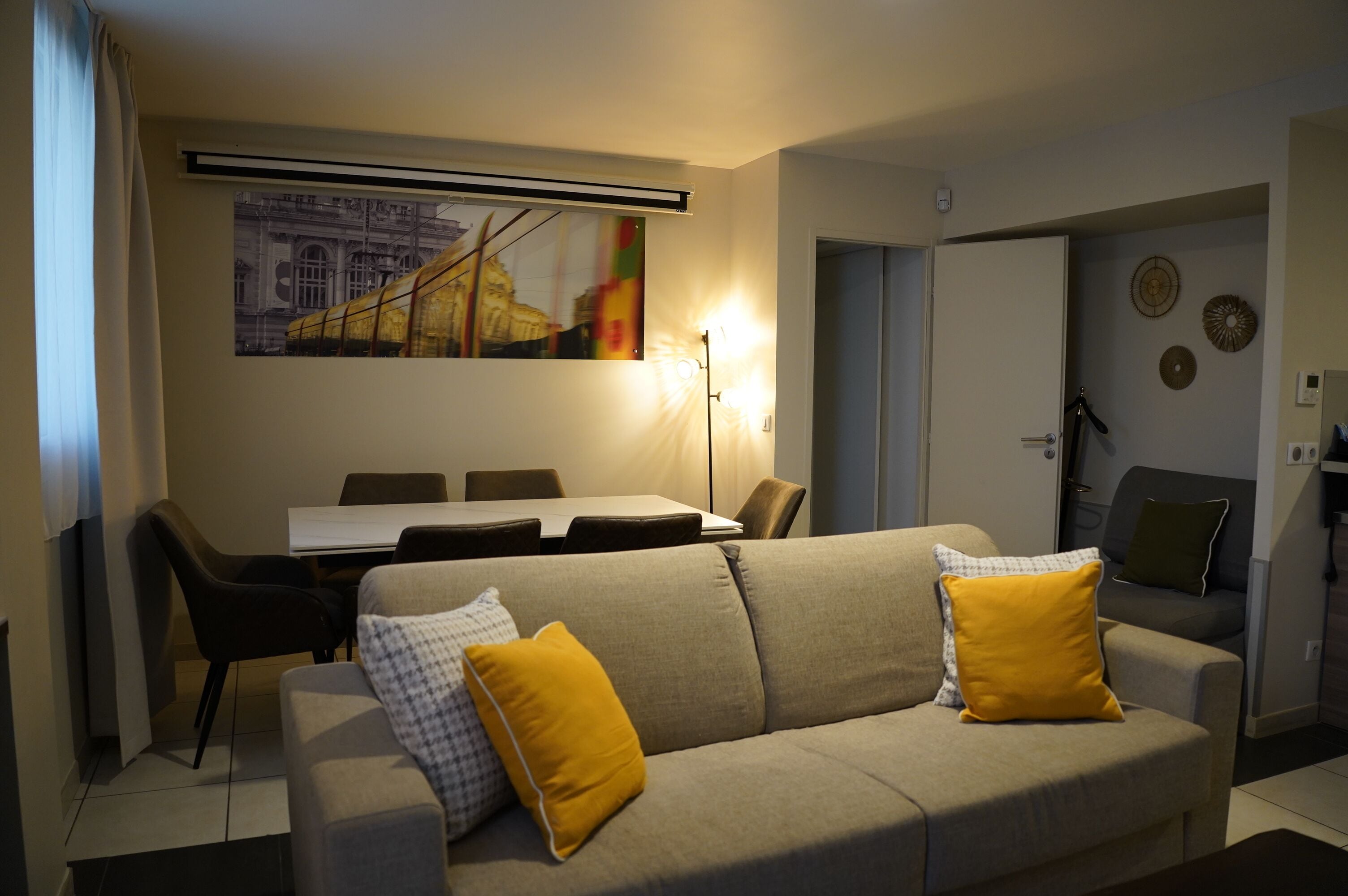 A living room with a sofa, a dining table with chairs, a painting on the wall, and a door leading to another room.