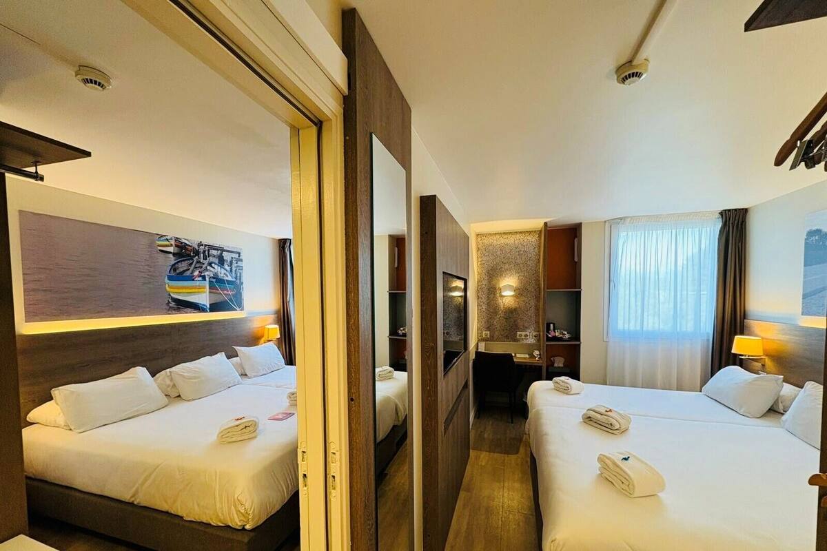 Habitación de hotel con dos camas, un escritorio, un espejo y vista a un barco a través de una ventana.