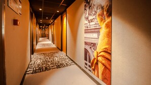 Ein Hotelgang mit Teppichmuster, einem großen Poster an der rechten Wand und einer Decke mit eingelassenen Leuchten.