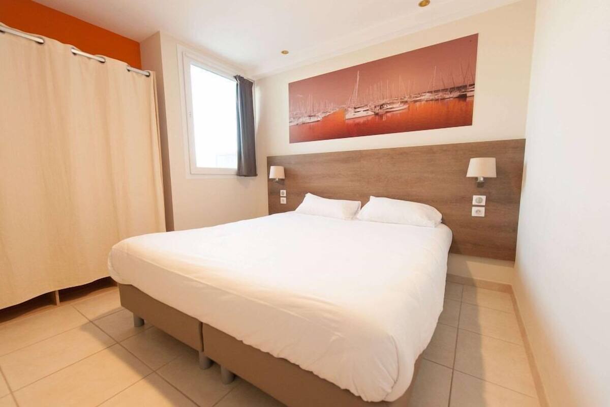 Habitación de hotel con una cama grande, una ventana con cortinas, una lámpara de pared y un cuadro que representa una escena portuaria.
