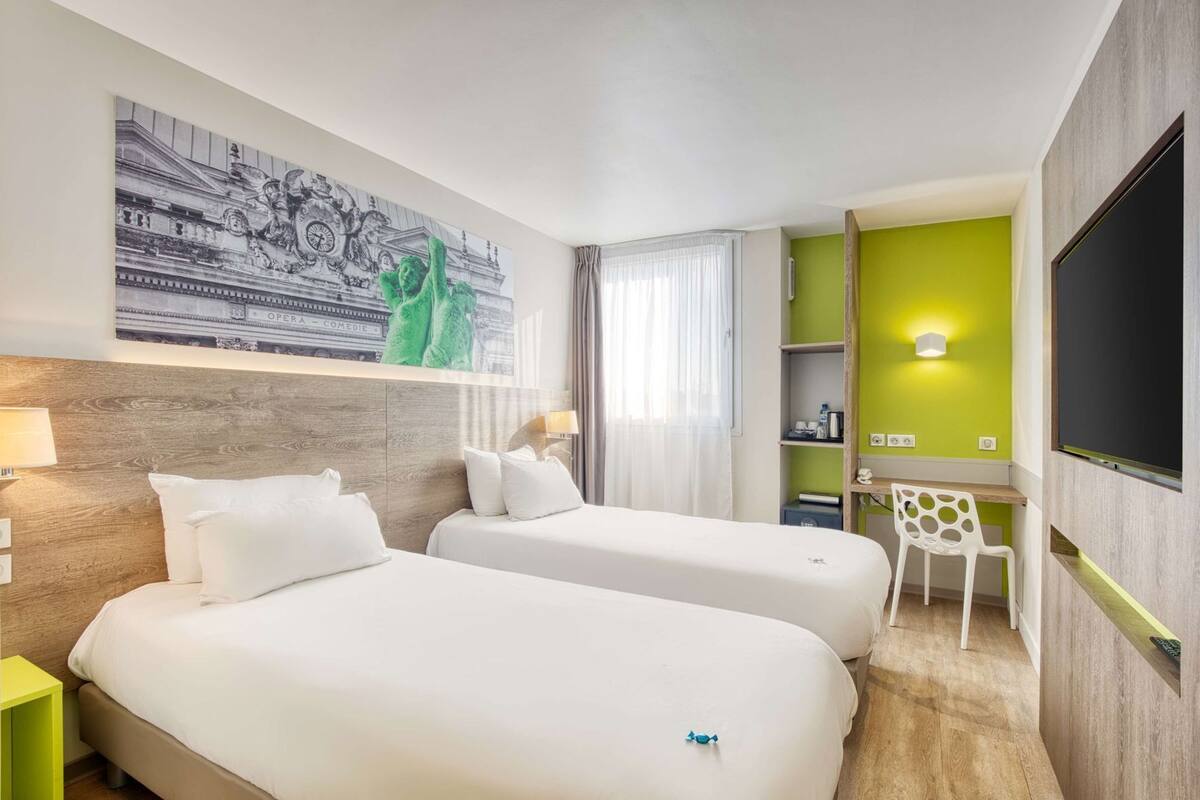 Habitación de hotel con dos camas, un escritorio, una silla, un televisor y una pared verde decorativa.