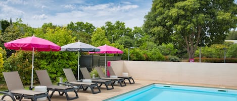 Ein Schwimmbecken mit Liegestühlen und rosa Sonnenschirmen, umgeben von viel Grün.