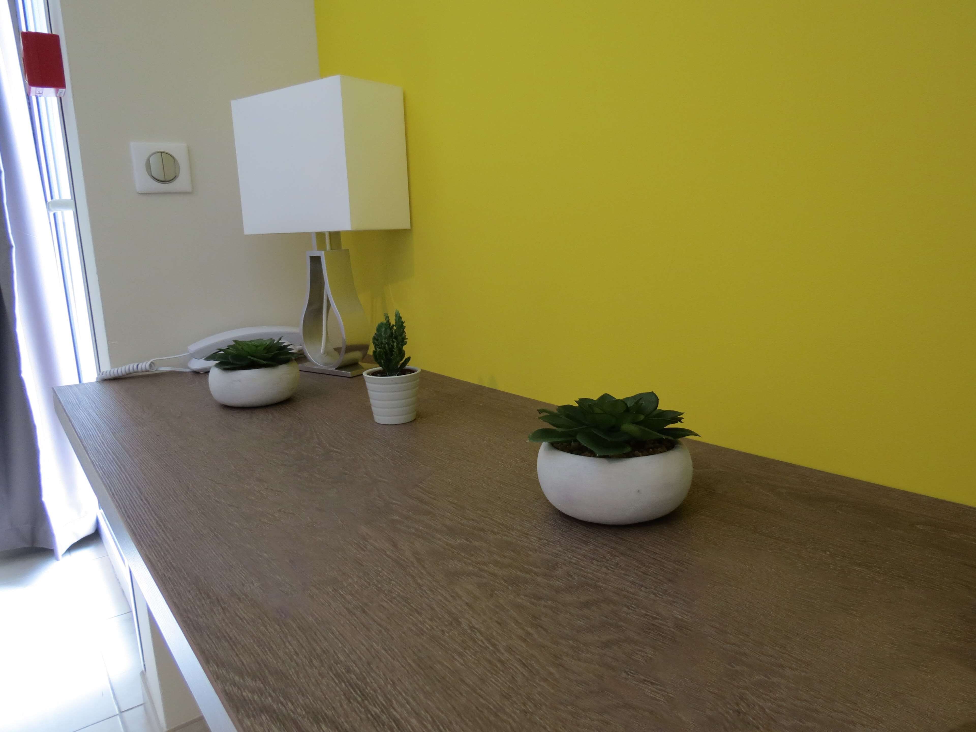 Een houten tafel met twee planten in potten en een witte lamp tegen een gele muur.