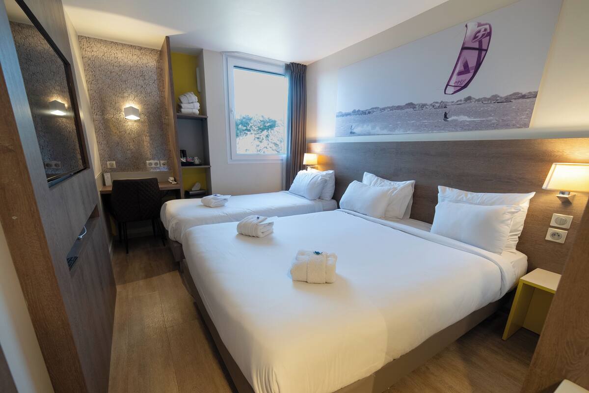 Habitación de hotel con dos camas, un escritorio, una silla, un espejo, una ventana y un televisor montado en la pared.