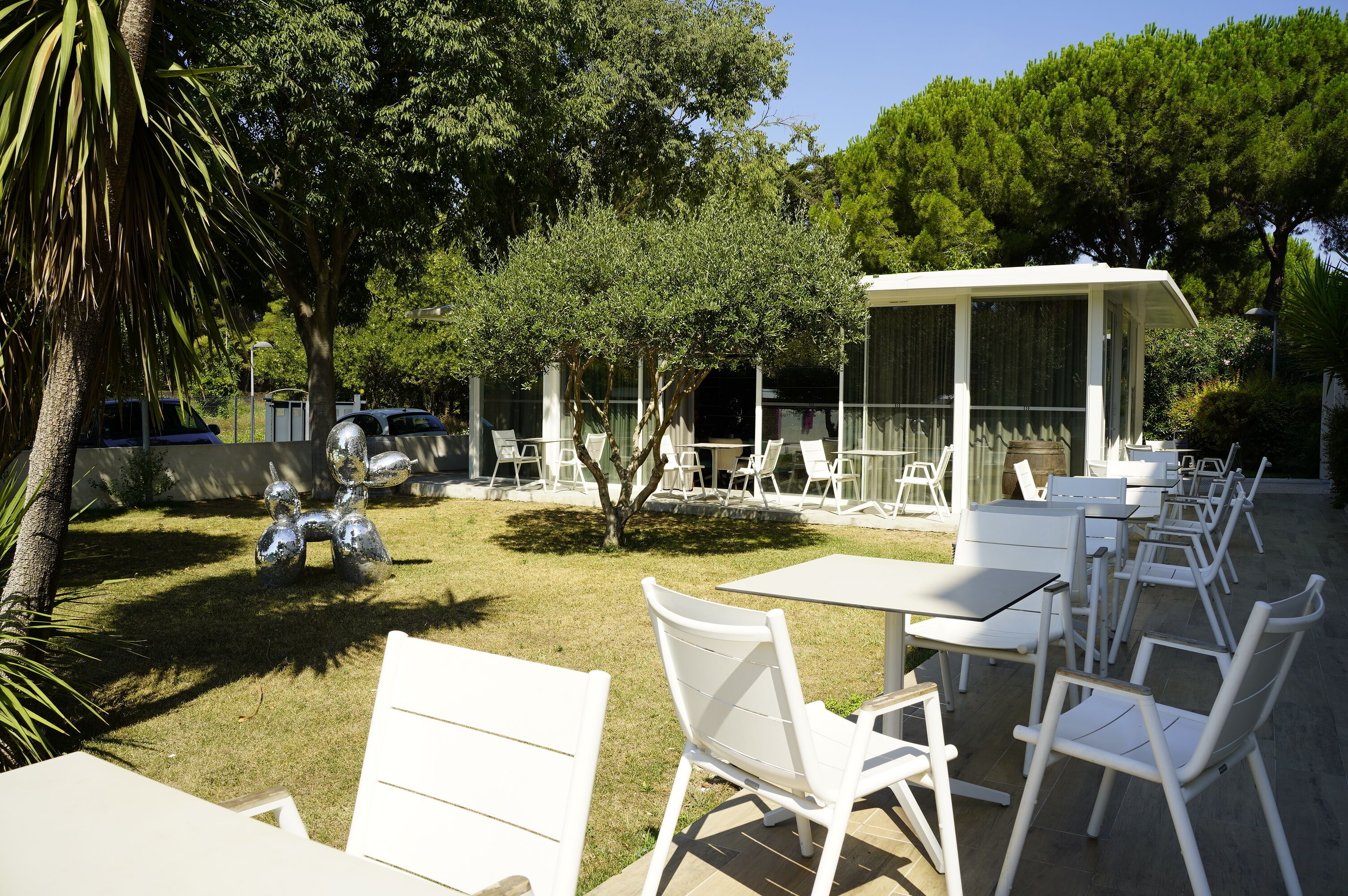 An outdoor seating area with white tables and chairs, surrounded by trees and a covered seating area.