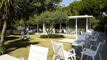 An outdoor seating area with white tables and chairs, surrounded by trees and a covered seating area.