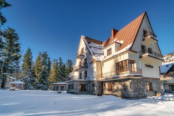A large, multi-story building with a stone facade and multiple balconies, surrounded by snow-covered trees.