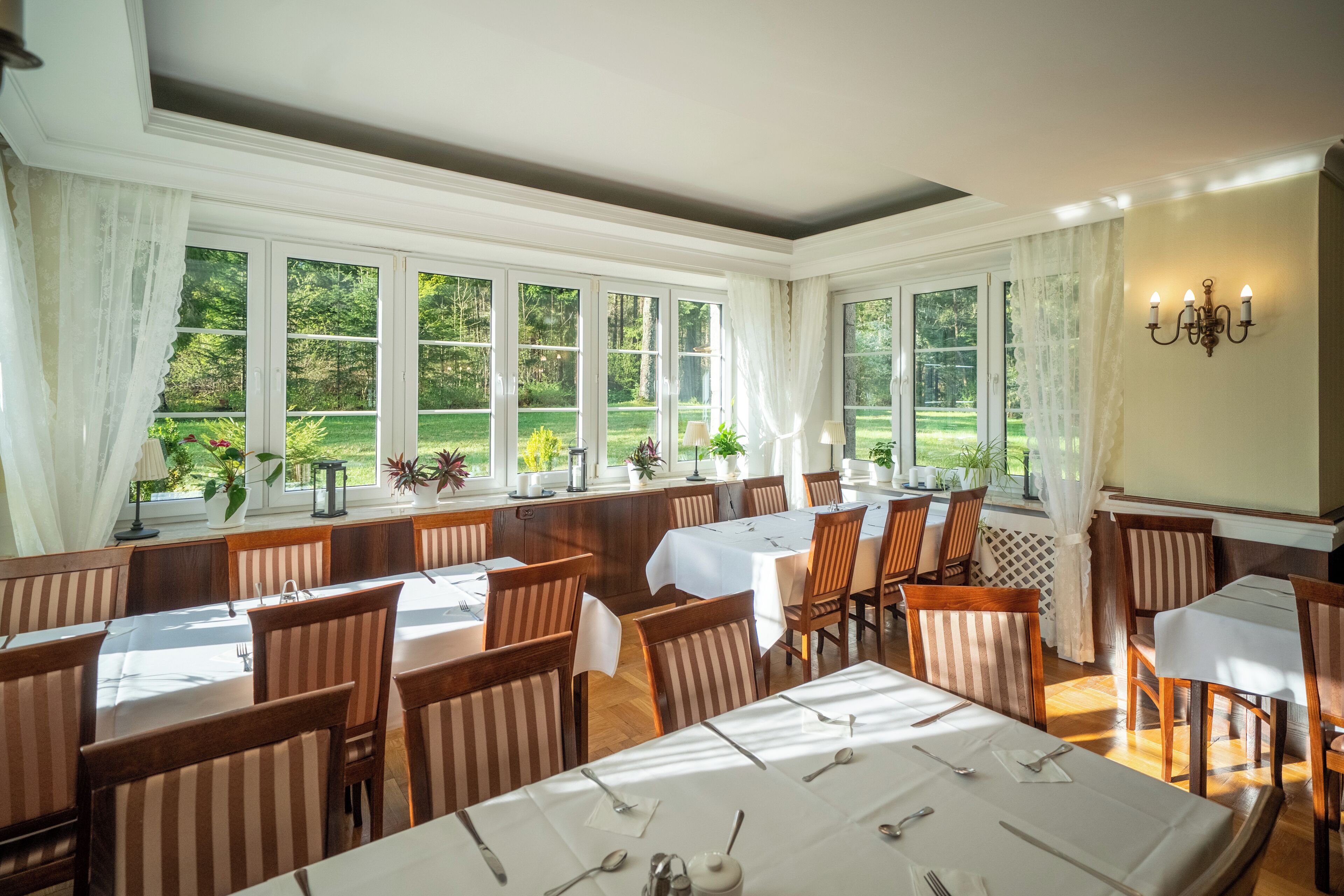A dining area with multiple tables and chairs, large windows with curtains, and a view of greenery outside.