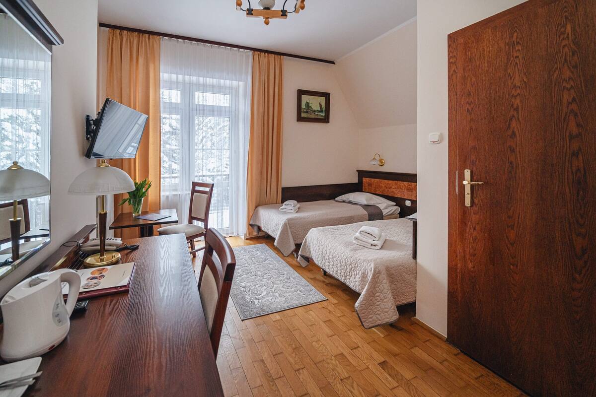 Une chambre d’hôtel avec deux lits, un bureau avec une lampe, un téléviseur fixé au mur et une porte en bois.