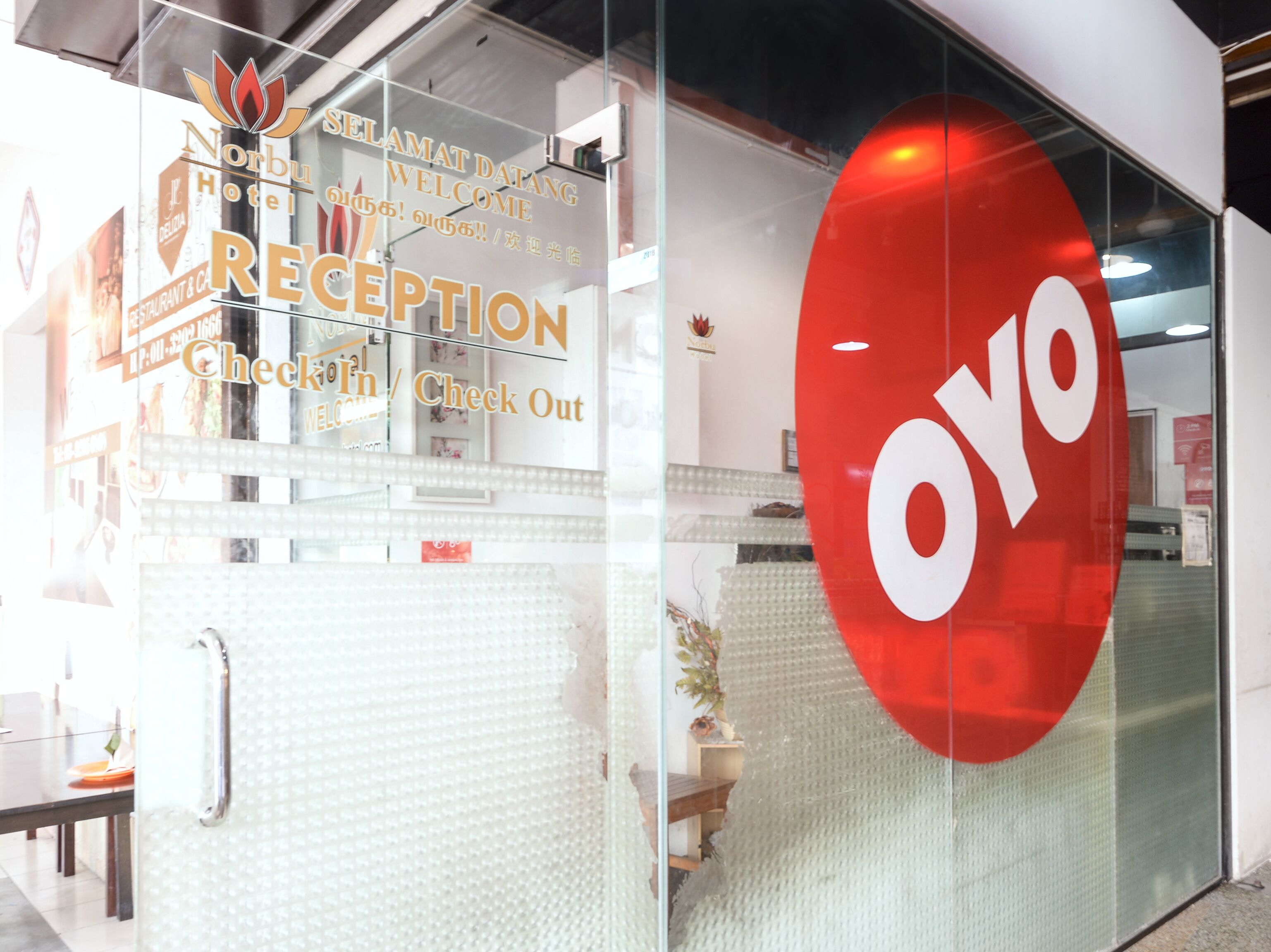 Una zona de recepción con un separador de vidrio, el logotipo rojo 'OYO' y un cartel que indica 'Registro de entrada / Registro de salida'.
