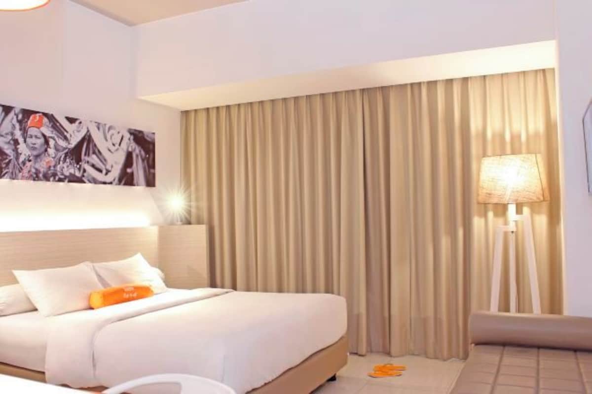 Una habitación de hotel con una cama, cabecero, un ventanal con cortinas, una lámpara de pared y un cuadro.