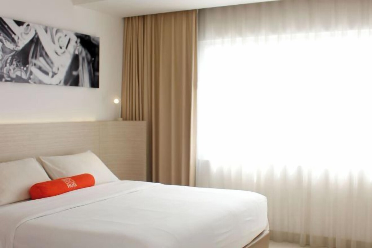 A hotel room with a bed, a large window with sheer curtains, and a wall-mounted artwork.