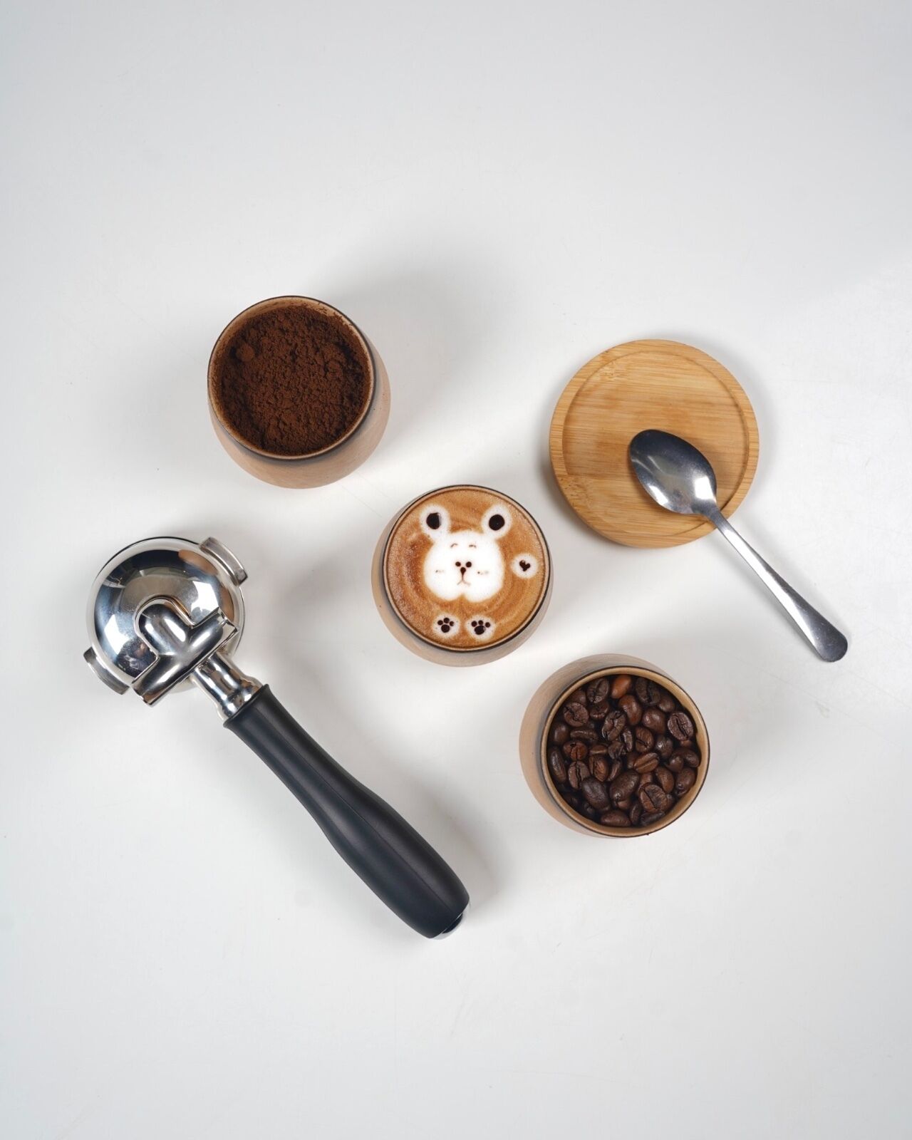 A coffee maker, a cup of coffee with latte art, a bowl of coffee beans, a scoop, and a container of coffee powder.